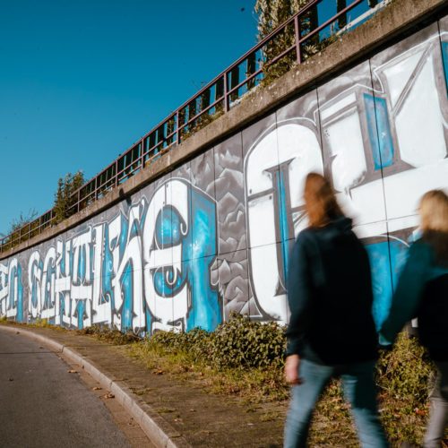 Zdjęcie przedstawia dwie osoby przed graffiti Schalke 04