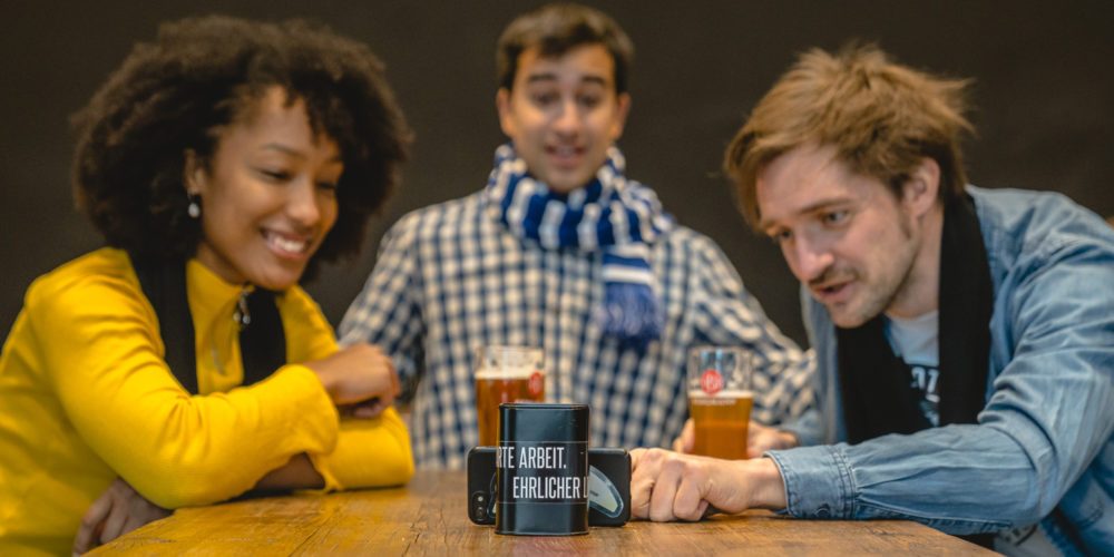 Das Bild zeigt drei Menschen, die ein Fußballspiel auf dem Handy verfolgen in der Stehbierhalle der Bergmann Brauerei Dortmund