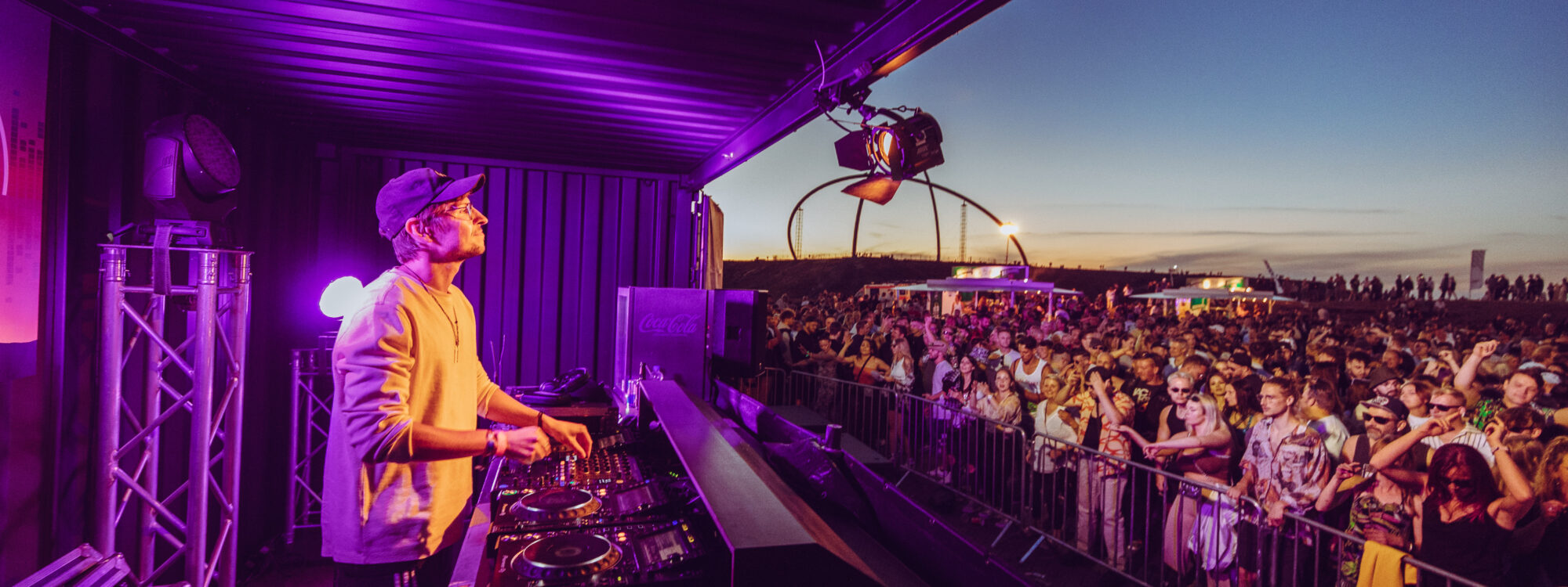 Ein DJ legt beim Picknick bei Sonnenuntergang auf einer Freilichtbühne Musik für eine große Menschenmenge auf, die tanzt und die Festivalatmosphäre bei bunter Beleuchtung genießt.