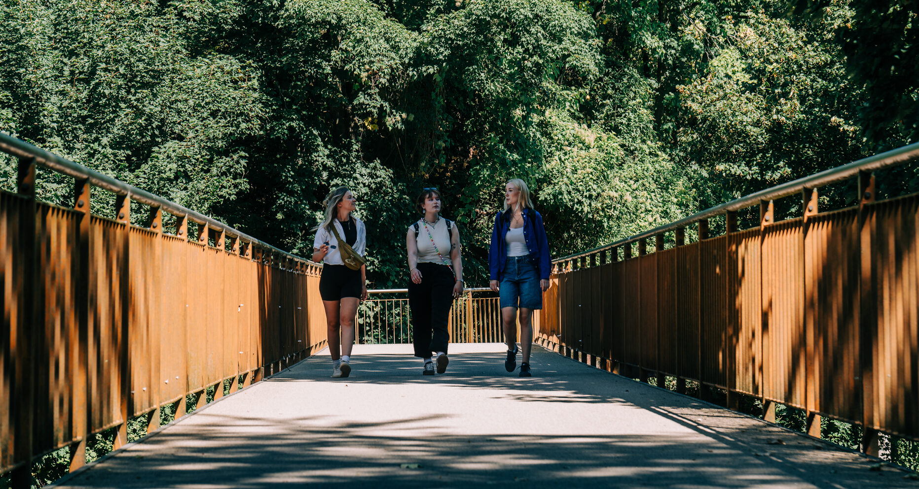 Drei Personen gehen nebeneinander auf einer sonnigen Fußgängerbrücke, die von üppigen grünen Bäumen umgeben ist, wobei Metallgeländer Schatten auf den Weg werfen.